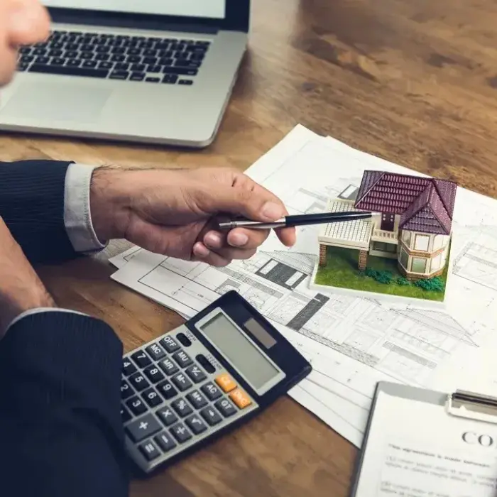 Experto de Gestión inversión calculando el valor de una propiedad, utilizando planos arquitectónicos y una maqueta de casa para un análisis detallado.