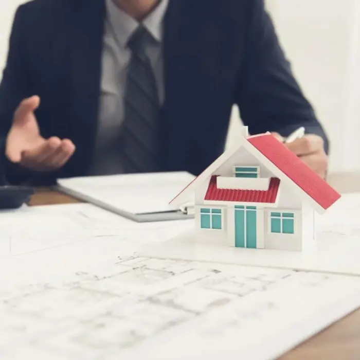 Un consultor de Gestión inversión explicando los detalles de un proyecto sobre planos arquitectónicos, con una casa a escala como modelo de referencia.